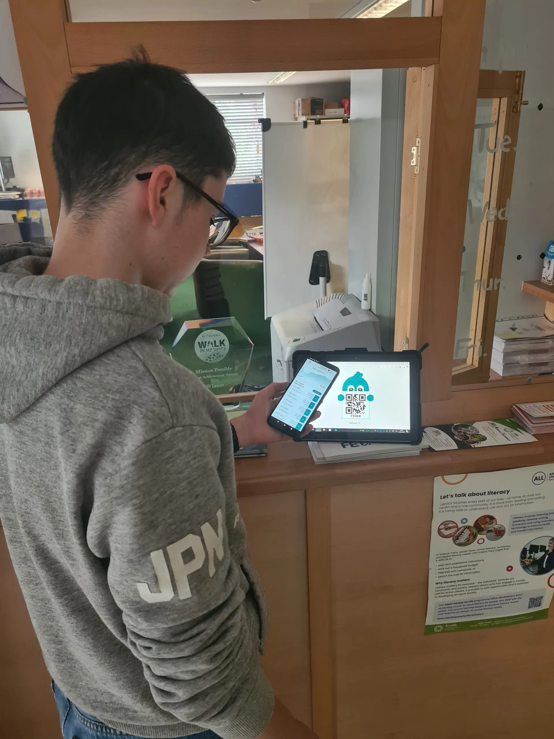 Kilkenny - student self checkin Male teenager standing with a phone in front of an iPad showing a QR code